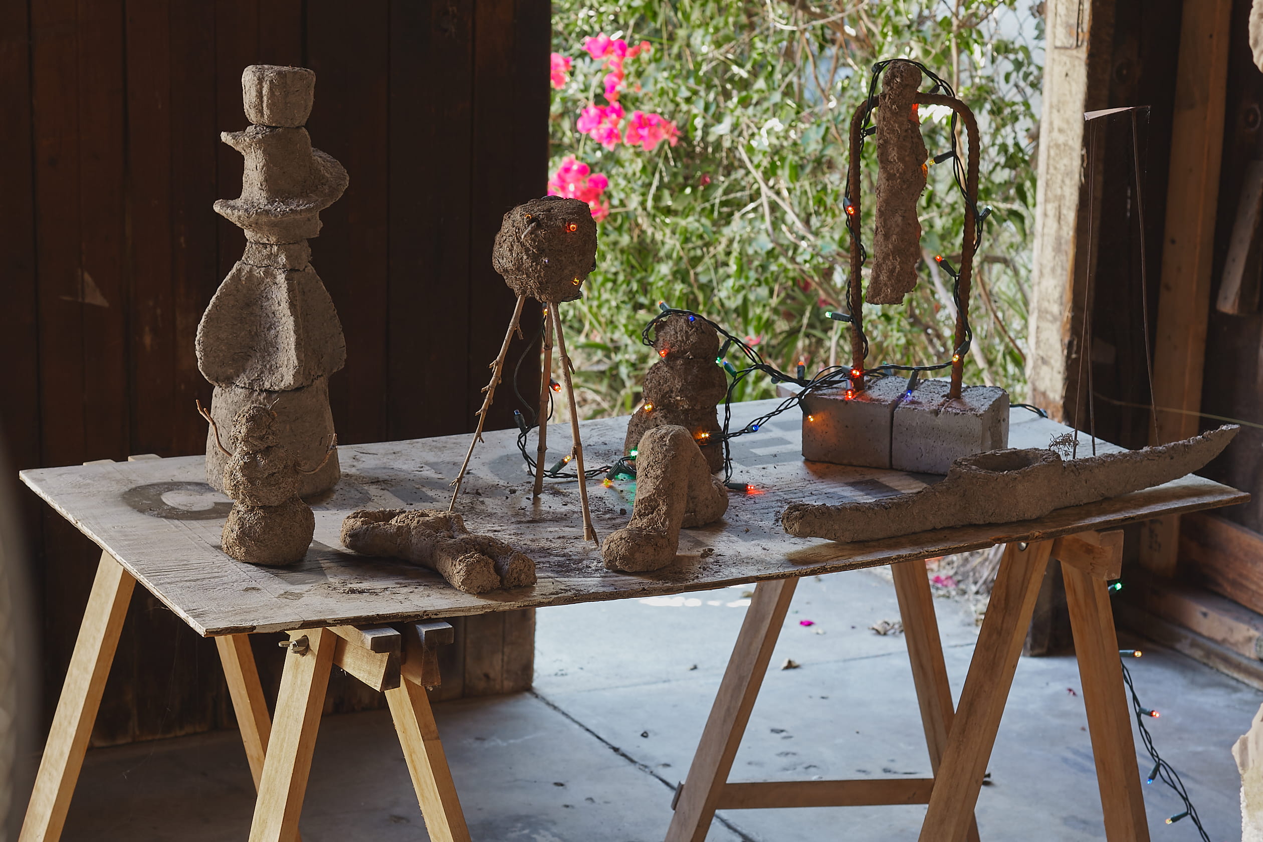 Untitled - A Sculpture By Brian Dario, featuring eight sculptures formed out of dirt, cinderblocks, rebar, and christmas lights on top of a sawhorse with a plank of wood used like a table, in front of an open door to a backyard with pink flowers.