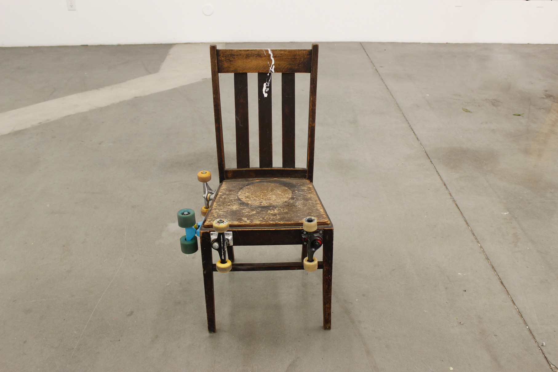 Wheelie Chair - A Sculpture by Brian Dario, An old wooden chair with four skateboard trucks, wheels still attatched, fastened to the front side, and the sitter's right-hand side.  There are no trucks fastened to the sitter's left-hand side